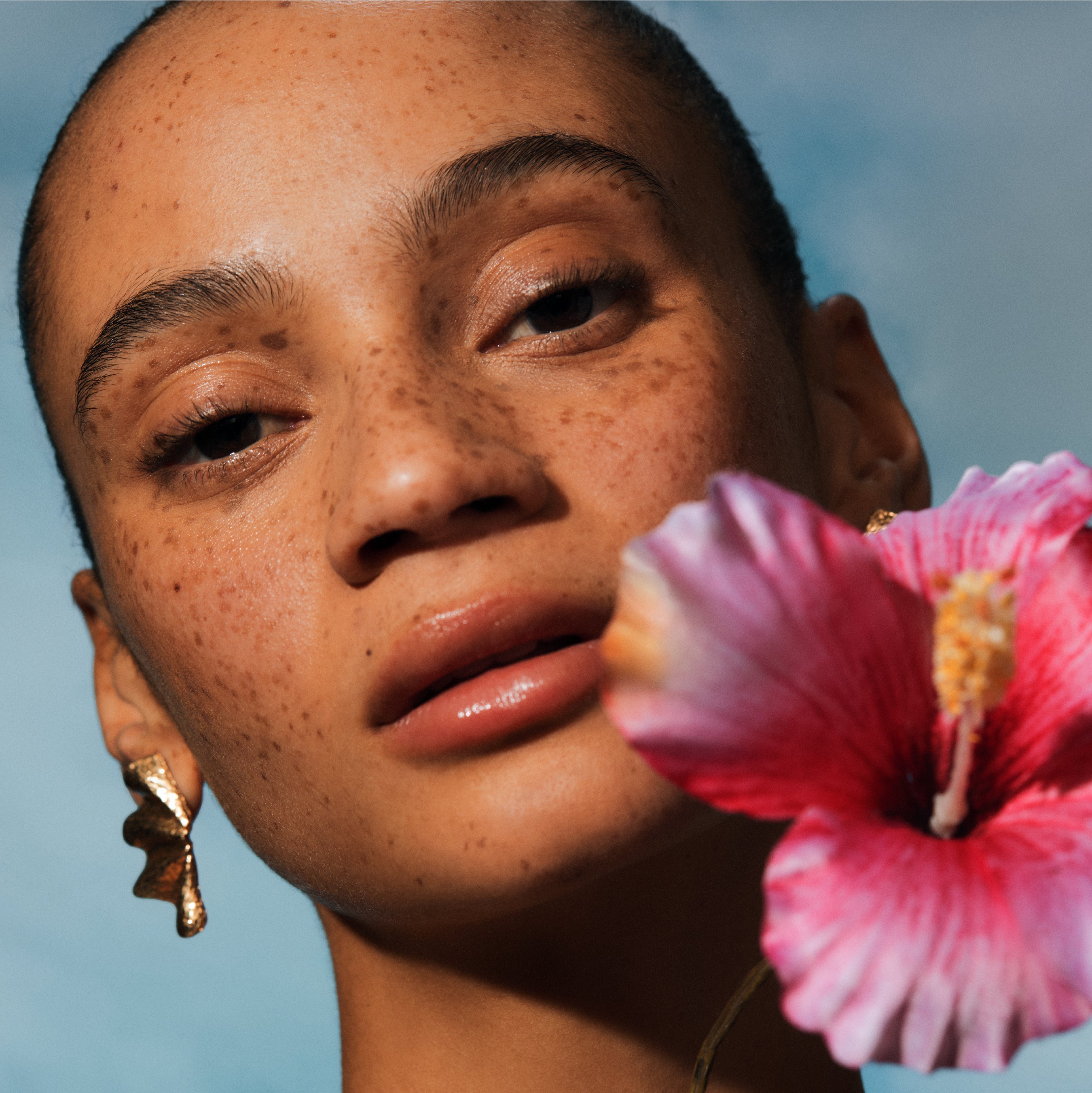 Close-up of a person with a pink Hibiscus flower in front of their face against a blue sky.